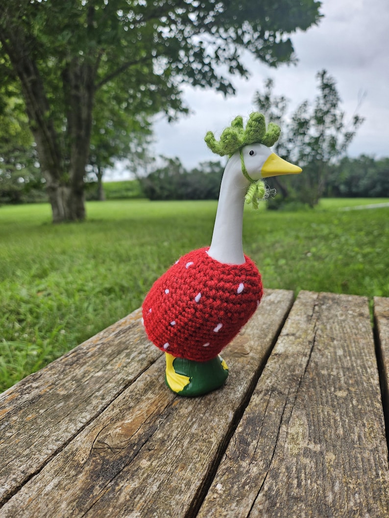 Puede incluir: Figura de ganso de cer&aacute;mica con cuello y cabeza blancos, pico amarillo y patas verdes y amarillas. El ganso lleva un disfraz de fresa de ganchillo rojo con detalles de semillas blancas y una corona de ganchillo verde. La figura est&aacute; sobre una superficie de madera.