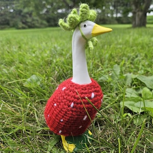 Puede incluir: Una figura de ganso de cer&aacute;mica, pintada de blanco con pico y patas amarillas, vestida con un disfraz de fresa roja de ganchillo con lunares blancos. Lleva un sombrero de hojas verdes de ganchillo, de pie en hierba verde.