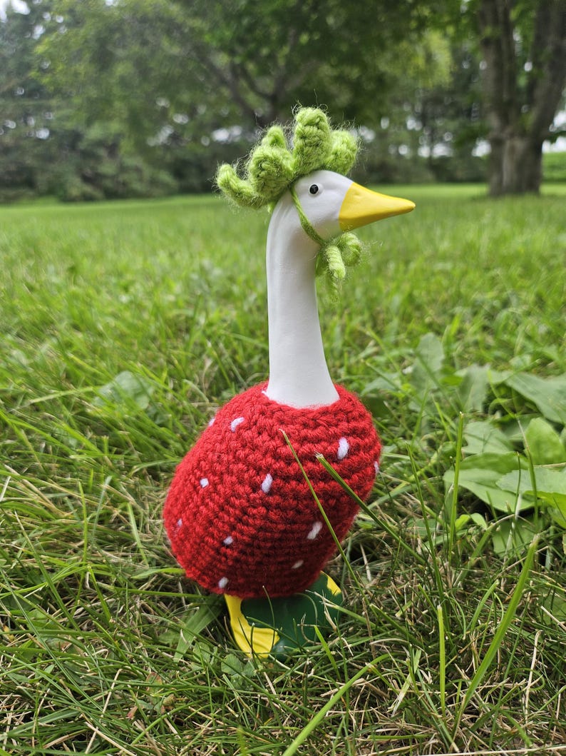 Puede incluir: Una figura de ganso de cer&aacute;mica, pintada de blanco con pico y patas amarillas, vestida con un disfraz de fresa roja de ganchillo con lunares blancos. Lleva un sombrero de hojas verdes de ganchillo, de pie en hierba verde.
