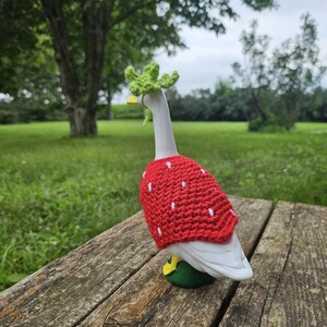 Puede incluir: Una figura de ganso de cer&aacute;mica con un disfraz de fresa roja de ganchillo, con detalles de semillas blancas y una corona de hojas verdes. El ganso tiene un pico amarillo y est&aacute; colocado sobre una superficie de madera con un fondo de hierba.