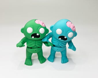 May include: Two small, 3D printed zombie figures. One is green with a pink brain, the other is blue with a pink brain. Both have white eyes and open mouths.