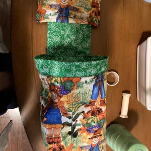 May include: A green and white fabric storage bin with a scarecrow pattern. The bin has a green fabric top with a white floral pattern and a green fabric bottom with a white floral pattern.