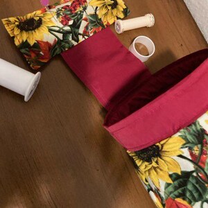 May include: A red fabric storage bag with a floral print featuring sunflowers and other flowers. The bag is lined with a pink fabric and has a flap with a floral print.