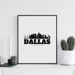 May include: Black and white framed print featuring the Dallas, Texas skyline silhouette. The word "DALLAS" is printed in bold, black letters, with "TEXAS" below it. The print is displayed on a white surface with a cactus and candle.