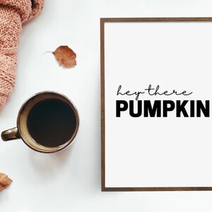 Könnte beinhalten: Gerahmter Druck mit dem Text "hey there PUMPKIN" in Schwarz. Eine Kaffeetasse, eine rosa Strickdecke und getrocknete Blätter sind auf einer weißen Oberfläche angeordnet und schaffen ein gemütliches Herbstthema.