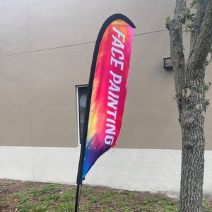May include: A colorful feather flag with the text "FACE PAINTING" printed on it. The flag is attached to a black pole and is displayed outdoors.