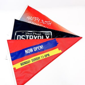 May include: Three triangular flags with different designs. The top flag is red and orange with the text "HAPPY 30TH BIRTHDAY". The middle flag is black with white text that reads "DAILY DRIVEN SINCE 2023 DSTVDIV". The bottom flag is red with a blue and yellow stripe and the text "NOW OPEN! MONDAY-SUNDAY 11-9PM".