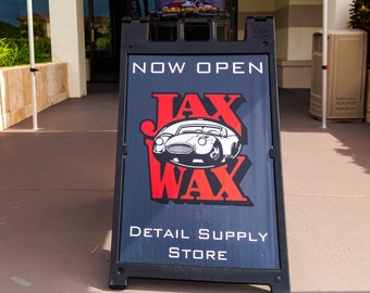 Puede incluir: Un letrero negro y rojo con las palabras "Now Open" y "Jax Wax Detail Supply Store". El letrero tiene una imagen de dibujos animados de un coche rojo sobre un fondo blanco.
