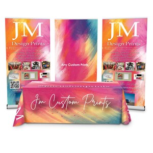 May include: A colorful table cover with the text "JM Custom Prints" in white script. The table cover is draped over a table with three colorful retractable banners behind it. The banners have the text "JM Design Prints" and "Any Custom Prints" on them.