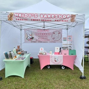 May include: A white tent with a pink and white table and a green table. The tent has a sign that says "Heidi's Bakeshop" with the words "Cookies, Sweet Breads, Pastries".