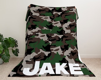 Può includere: Una coperta in pile con motivo mimetico verde, marrone e grigio con il nome "JAKE" stampato in bianco sul fondo.