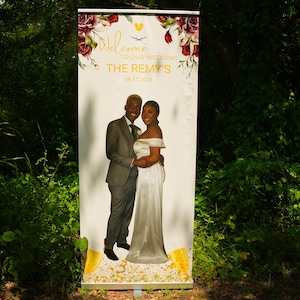 May include: A white wedding sign with floral designs and a photo of a couple. The sign reads "Welcome to our wedding The Remy's 08.17.2024".