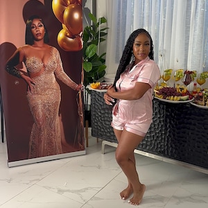 May include: A woman wearing a pink satin pajama set with short sleeves and shorts. She is standing in a room with white tile floors and a large poster behind her. The poster features a woman in a gold sequined dress.