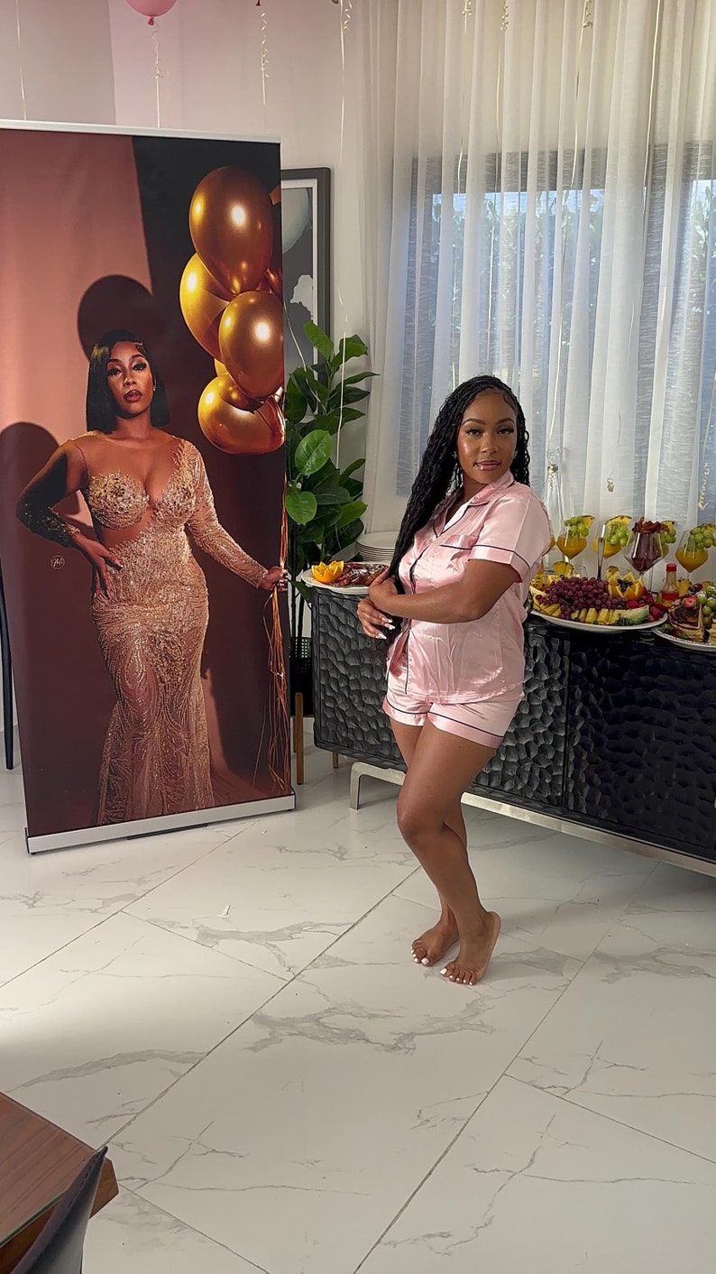 May include: A woman wearing a pink satin pajama set with short sleeves and shorts. She is standing in a room with white tile floors and a large poster behind her. The poster features a woman in a gold sequined dress.
