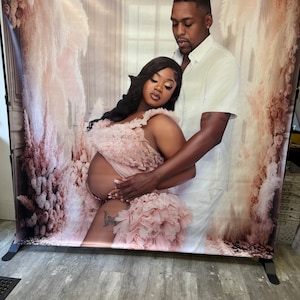 May include: A couple poses for a maternity photo shoot. The woman is wearing a pink ruffled dress and the man is wearing a white shirt. The background is a white backdrop with floral accents.