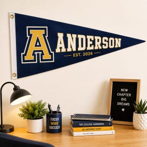 May include: A triangular navy blue felt pennant featuring a large yellow letter A with a white outline, the text ANDERSON in white, and the text EST. 2026 in yellow. The pennant has a white vertical strip on the left side with two brass-colored metal grommets.