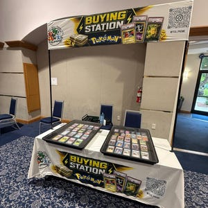 May include: A trade show booth featuring a white table cover with the text BUYING STATION and Pokémon branding. Two black display trays filled with collectible trading cards sit on the table. A matching banner hangs above with a QR code.