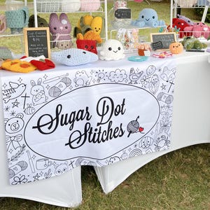 May include: A white table cloth with a black and white design featuring a variety of cute illustrations. The table cloth has a large oval with the text "Sugar Dot Stitches" in black. The table is covered with crocheted items, including a variety of animals, flowers, and other objects. The table is set up at a craft fair.