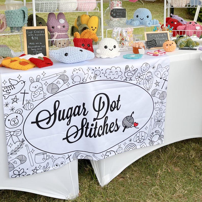 May include: A white table cloth with a black and white design featuring a variety of cute illustrations. The table cloth has a large oval with the text "Sugar Dot Stitches" in black. The table is covered with crocheted items, including a variety of animals, flowers, and other objects. The table is set up at a craft fair.