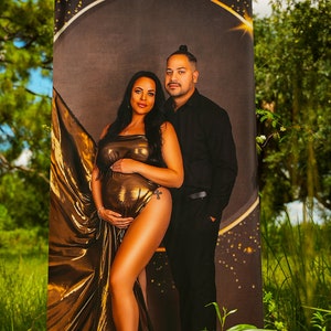 May include: A couple poses for a maternity photo shoot. The woman is wearing a gold, flowing dress and the man is wearing a black shirt and pants. The background is a dark brown with gold glitter.