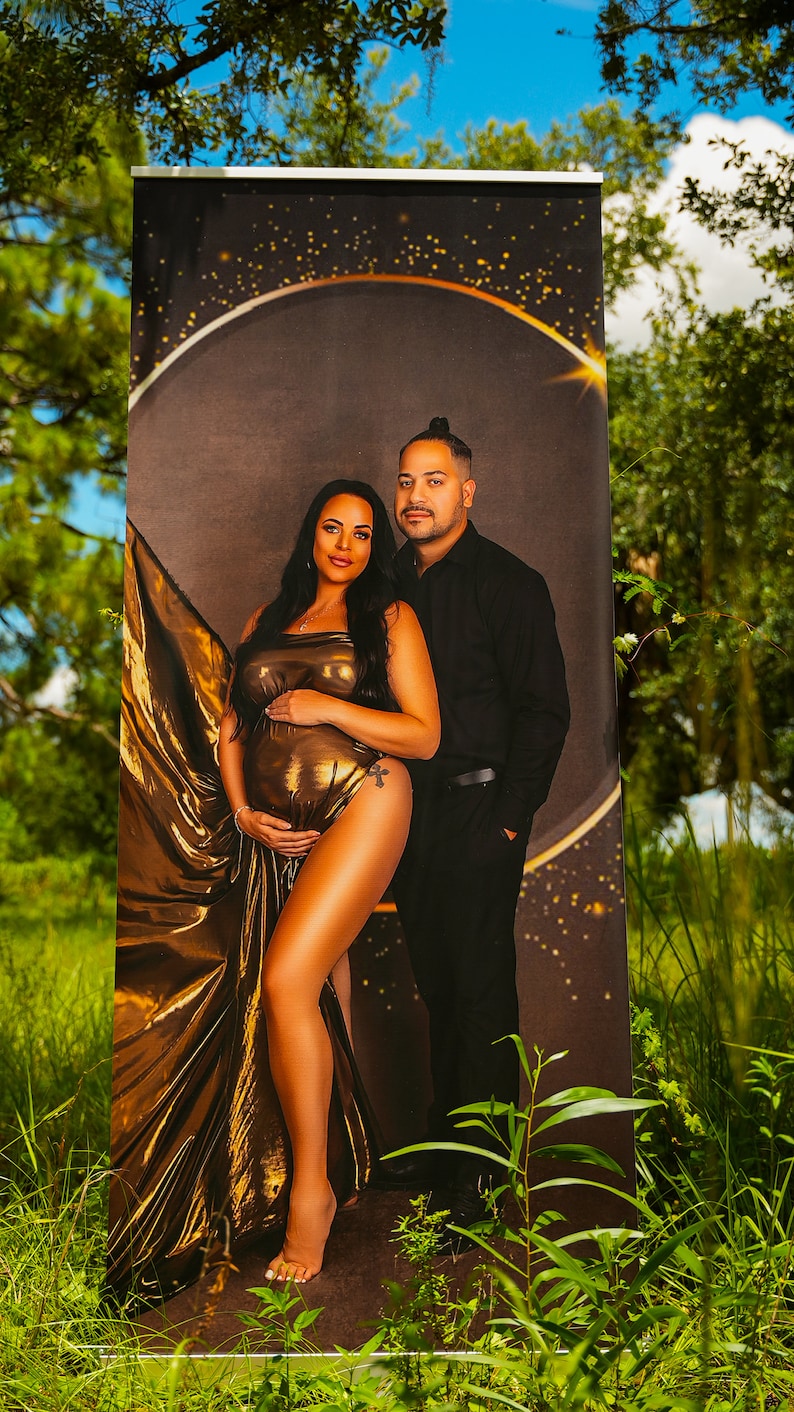 May include: A couple poses for a maternity photo shoot. The woman is wearing a gold, flowing dress and the man is wearing a black shirt and pants. The background is a dark brown with gold glitter.