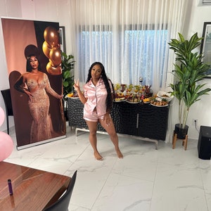 May include: A woman in a pink satin pajama set poses in front of a black and silver dresser. The room is decorated with pink and gold balloons.