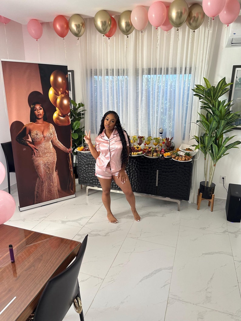 May include: A woman in a pink satin pajama set poses in front of a black and silver dresser. The room is decorated with pink and gold balloons.