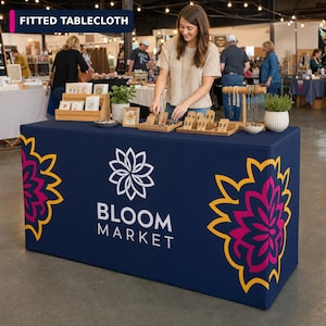 May include: A navy blue fitted tablecloth featuring a white Bloom Market logo and a stylized flower graphic in pink and yellow on each side. The fabric covers a rectangular table, displaying a clean, smooth surface with a professional appearance.