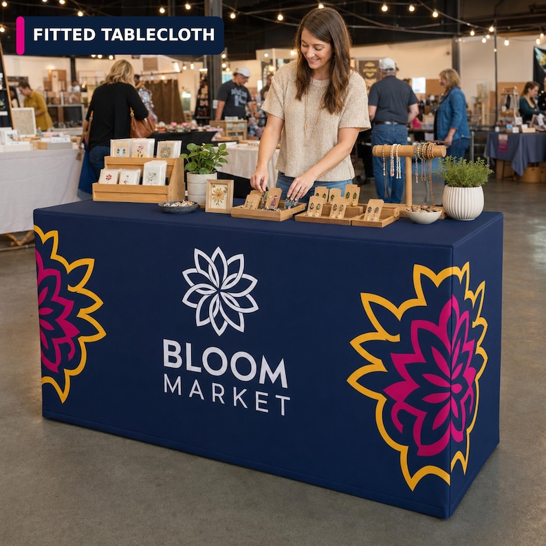 May include: A navy blue fitted tablecloth featuring a white Bloom Market logo and a stylized flower graphic in pink and yellow on each side. The fabric covers a rectangular table, displaying a clean, smooth surface with a professional appearance.