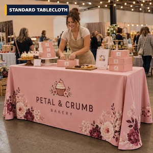May include: A rectangular pink tablecloth featuring a floral pattern of roses and leaves along the bottom edge. The center displays a graphic of a cupcake surrounded by flowers and the text Petal & Crumb Bakery in dark brown lettering.