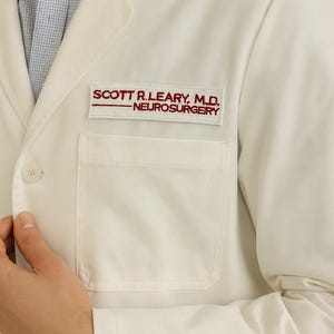 May include: A white medical lab coat featuring a pocket and the embroidered name "SCOTT R. LEARY, M.D. - NEUROSURGERY" in red thread. The coat is buttoned and worn over a light blue patterned shirt.