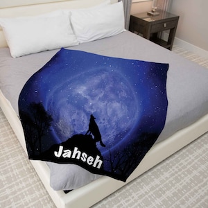 Puede incluir: Una manta de franela azul y negra con un lobo aullando a la luna. La luna est&aacute; llena y rodeada de estrellas. La manta tiene el nombre "Jahseh" impreso en ella.