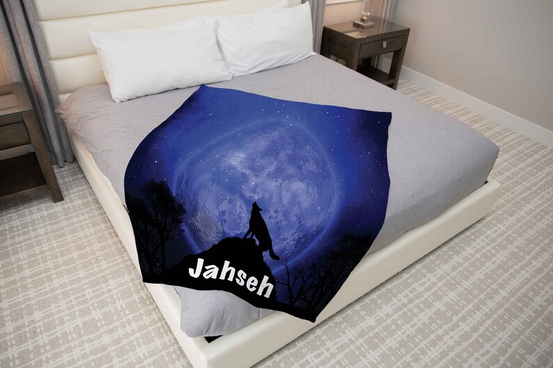 Puede incluir: Una manta de franela azul y negra con un lobo aullando a la luna. La luna est&aacute; llena y rodeada de estrellas. La manta tiene el nombre "Jahseh" impreso en ella.