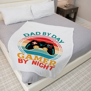 May include: White blanket with a retro design featuring a black video game controller and the text "DAD BY DAY GAMER BY NIGHT" in teal, red, orange, and yellow. The blanket is folded on a bed with gray sheets and pillows.