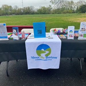May include: A white table with a black tablecloth and a white table runner with the logo "Above the Horizon Consulting, LLC" and a blue and green logo of a river winding through a green hill. The table is set up with brochures, pens, candy, and other promotional materials.