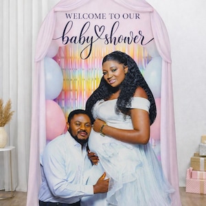 Puede incluir: Un tel&oacute;n de fondo de baby shower con una pareja. El tel&oacute;n de fondo tiene un arco rosa con las palabras "WELCOME TO OUR baby shower" en escritura. Se ven globos rosas y azules. La mujer lleva un vestido blanco y el hombre una camisa azul claro.