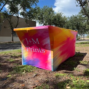 May include: A rectangular stretch table cover featuring a vibrant watercolor-style print in shades of pink, orange, and yellow. The front panel displays the white text J+M Prints in a clean, sans-serif font against the colorful background.