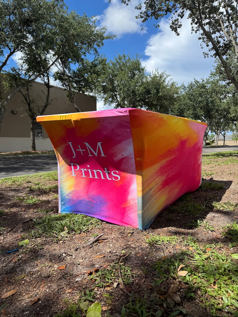 May include: A rectangular stretch table cover featuring a vibrant watercolor-style print in shades of pink, orange, and yellow. The front panel displays the white text J+M Prints in a clean, sans-serif font against the colorful background.