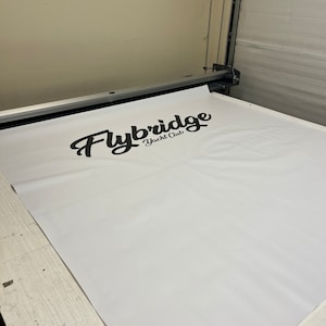 Pu&ograve; includere: Uno striscione bianco con il testo "Flybridge Yacht Club" stampato in corsivo nero. Lo striscione viene inserito in una grande macchina da taglio.