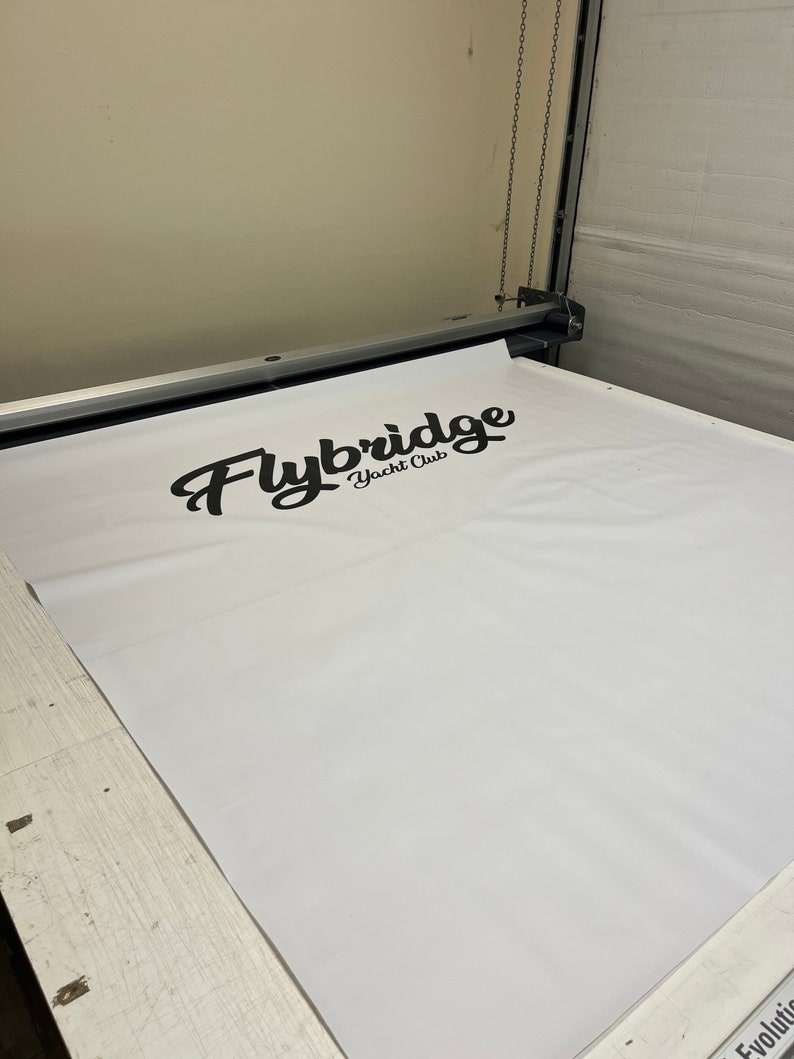 Pu&ograve; includere: Uno striscione bianco con il testo "Flybridge Yacht Club" stampato in corsivo nero. Lo striscione viene inserito in una grande macchina da taglio.