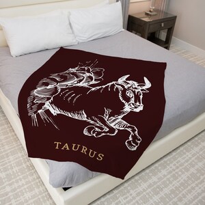 Könnte beinhalten: Eine weinrote Kuscheldecke mit einer weißen Illustration eines Stiers und dem Wort "TAURUS" in goldenen Buchstaben.