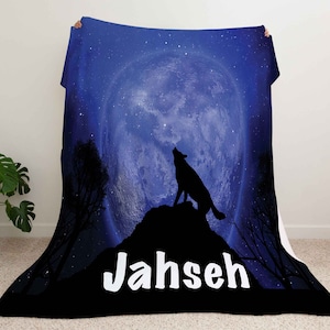 Puede incluir: Una manta de franela azul y negra con la silueta de un lobo aullando a la luna. La luna est&aacute; llena y rodeada de estrellas. La manta tiene el nombre "Jahseh" impreso en letras blancas.
