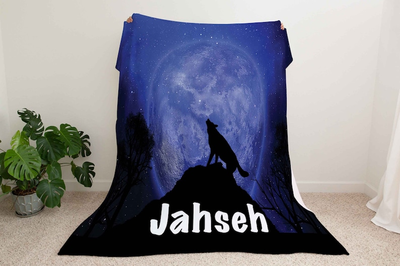 Puede incluir: Una manta de franela azul y negra con la silueta de un lobo aullando a la luna. La luna est&aacute; llena y rodeada de estrellas. La manta tiene el nombre "Jahseh" impreso en letras blancas.