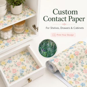May include: Custom contact paper with a floral pattern of pink, yellow, and blue flowers on a cream background, applied to white cabinet shelves and a drawer. A roll of the same paper sits nearby, alongside text reading Custom Contact Paper.