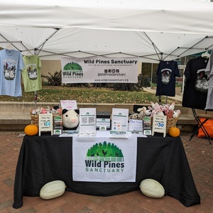 Puede incluir: Una mesa negra cubierta con un mantel negro con un logotipo blanco que dice "Wild Pines Sanctuary". Hay varios artículos en la mesa, incluyendo un cerdo de peluche, una calabaza, un letrero que dice "¡Llévatelo a casa! 1,50 €", y un letrero que dice "¡Cuida la granja con una donación de 30 €!" También hay varias camisetas colgadas en un estante detrás de la mesa.