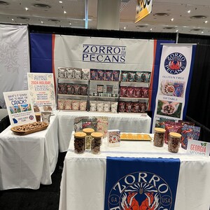 May include: A trade show booth with a white tablecloth and a blue and red banner that says "Zorro Pecans GA USA". There are jars of pecans on the table and a sign that says "2024 Holiday Limited Edition".