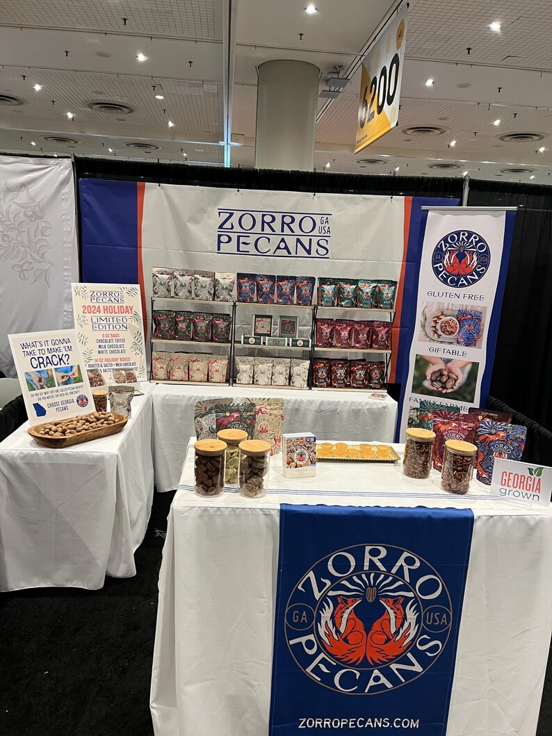 May include: A trade show booth with a white tablecloth and a blue and red banner that says "Zorro Pecans GA USA". There are jars of pecans on the table and a sign that says "2024 Holiday Limited Edition".