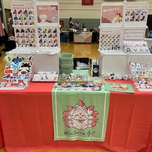May include: A table top display of handmade goods at a craft fair. The table is covered in a red tablecloth with a green banner in the center. The banner features a cartoon fox with the text "Winterra Art". The table is decorated with various items for sale, including keychains, enamel pins, and stickers. The items are displayed on shelves and in clear acrylic boxes. The text "Keychains 1 for $10, 3 for $25" is visible on the left side of the table. The text "Enamel Pins 1 for $5, 3 for $25" is visible on the right side of the table.