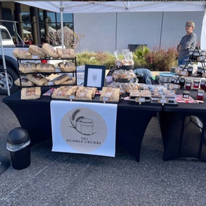 May include: A black table covered with a black tablecloth displays a variety of baked goods and jars of jam. The table has a sign that reads "Terri's Humble Crumbs".