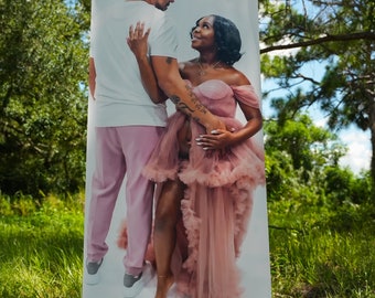 May include: A couple poses for a photo, the woman in a pink tulle dress and the man in a white shirt and pink pants. The photo is printed on a white banner.